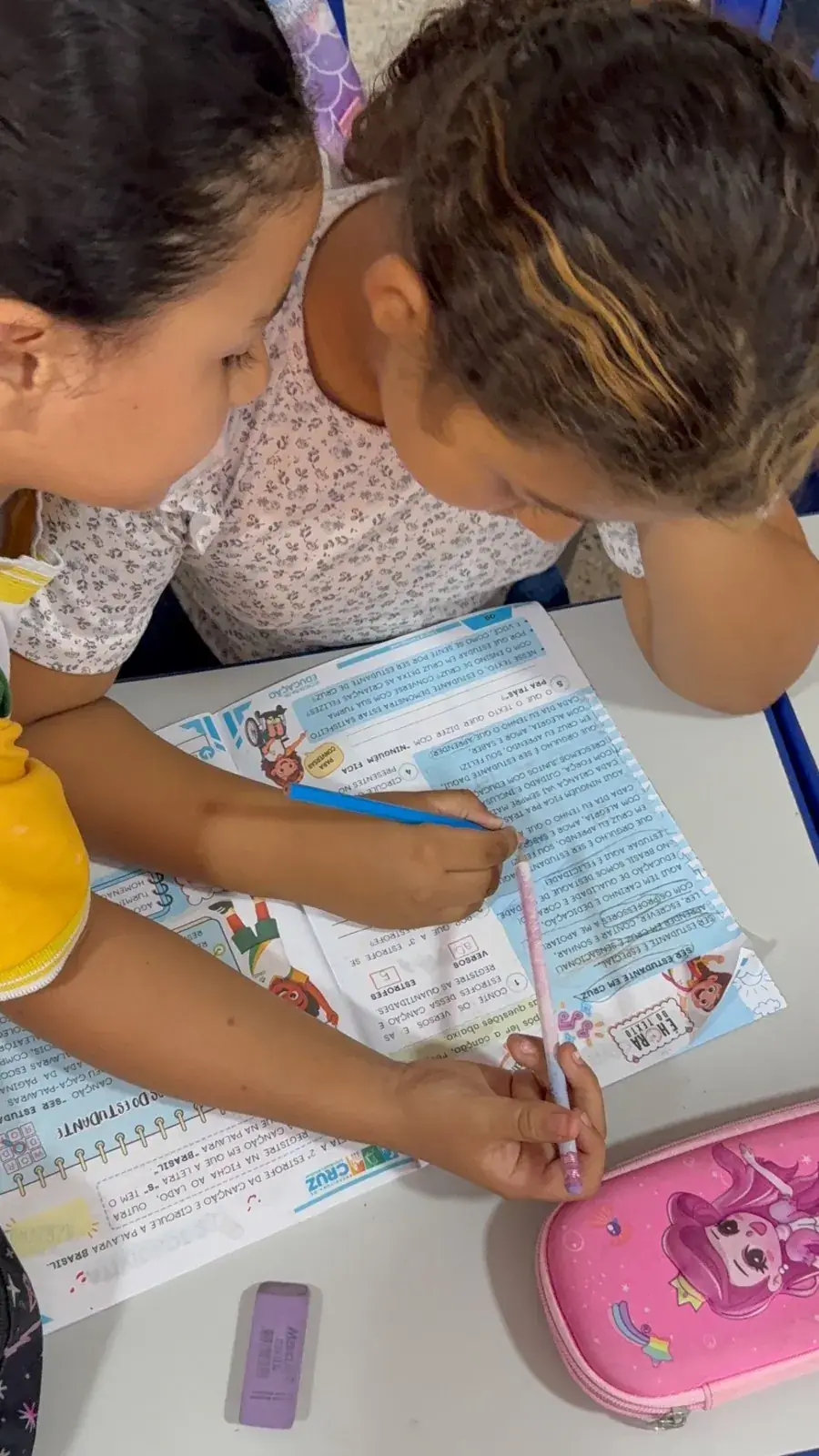 Estudante escreve no caderno de atividades durante a aula, com o material aberto sobre a carteira