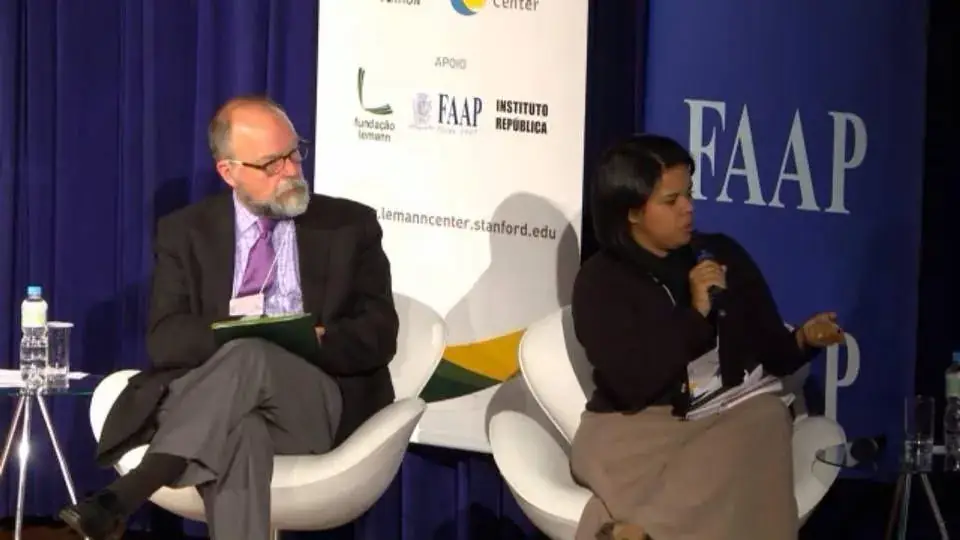 Janaína Barros fala ao microfone em painel de debate, com painel de apoio do Lemann Center e da FAAP ao fundo